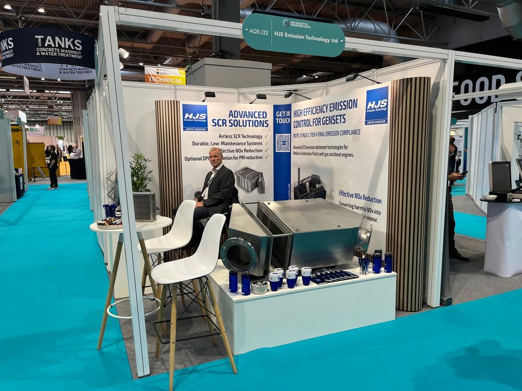 AQE Show image 3 - HJS UK Man in a suit seated at an HJS Emission Technology booth, with banners about SCR solutions and a metallic processing unit nearby.
